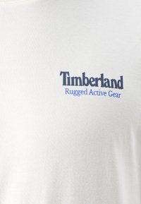 White cotton t-shirt featuring "Timberland" logo in black and "Rugged Active Gear" in blue. Smooth texture with a crew neck design.