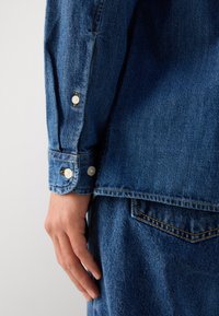 Blue denim long-sleeve shirt featuring white buttons on the cuffs, visible stitching, and a straight hemline with a casual fit.