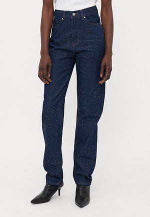 Mørkeblå denimjeans med høy midje, lette slitasjedetaljer og glitrende dekorasjoner. Rett ben passform, kombinert med svarte hæler.