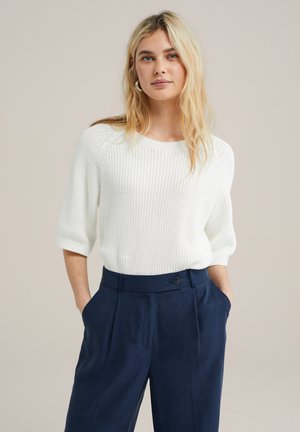 Pull en tricot blanc avec des manches courtes retroussées, associé à un pantalon taille haute bleu marine avec une fermeture par bouton à l'avant et des poches.