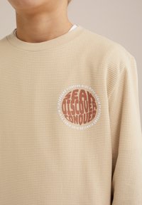 Pull long beige avec un motif texturé. Il présente un graphisme circulaire marron et blanc avec l'inscription "CHERCER DÉCOUVRIR CONQUÉRIR."