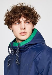 Giacca zip-up blu navy con colletto verde. Presenta coulisse e un logo discreto. Leggera, con una texture liscia e dal design sportivo.