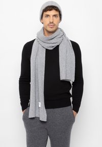 Harmaa ribattuja huivi ja siihen sopiva pipo, jotka on puettu mustan neuletakin päälle. Huivi on pitkä ja pehmeä, ja sen päässä on näkyvästi etiketti.