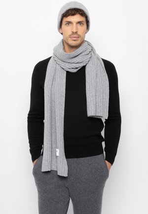 Grau gestreifter Schal und passende Mütze, getragen über einem schwarzen Pullover. Der Schal ist lang und weich, mit einem Etikett, das am Ende sichtbar ist.