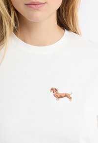 T-shirt bianca in cotone con un piccolo bassotto marrone ricamato sul petto a sinistra, con cuciture dettagliate e un'espressione giocosa.
