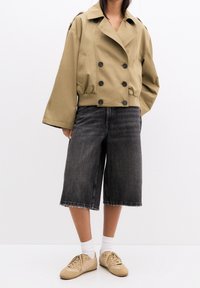 Kvinde iført en cropped beige dobbeltbrystet jakke, sorte brede denimshorts, hvide sokker og beige sneakers, som står foran en ensfarvet hvid baggrund.