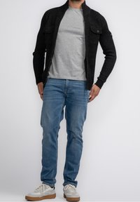 Mörkgrå zip-up-tröja med två bröstfickor, buren över en ljusgrå t-shirt. Ljusblå jeans och beige sneakers fulländar outfiten.