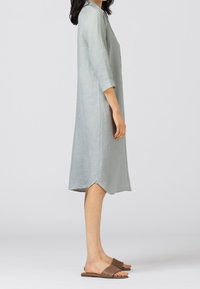 Kvinna klädd i en ljusgrå knälång klänning med knappar och 3/4-långa ärmar, samt bruna vävda platta sandaler, stående i profil mot en grå bakgrund.