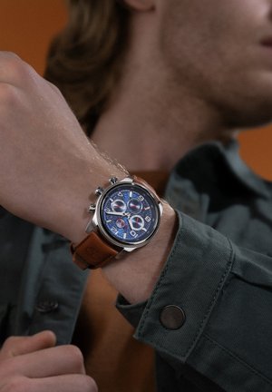 Orologio cronografo con quadrante blu, cassa in acciaio inossidabile con finitura argentata e cinturino in pelle marrone. Presenta tre quadranti secondari e numeri ben visibili.