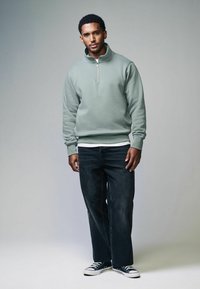 Felpa zipata verde menta con colletto alto, maniche lunghe e orlo a coste, abbinata a jeans dritti in lavaggio scuro e sneakers nere.