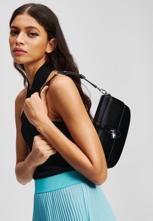 Sac à main en faux cuir noir avec une forme structurée, des accessoires en argent et une bandoulière amovible. Le modèle porte un haut noir à côtes et une jupe plissée turquoise.