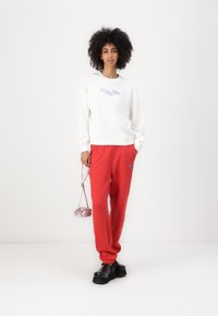 Hanorac alb cu logo albastru, pantaloni de trening roșii cu text albastru, pantofi cu platformă negri și o geantă mică roz. Materiale netede, croială lejeră.