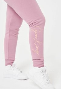 Růžové bavlněné tepláky s ribbovanými manžetami a výraznou béžovou výšivkou "GymKing" na spodní části nohy, doplněné bílými teniskami.