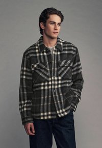 Chaqueta de franela a cuadros en blanco y negro con dos bolsillos en el pecho, cierre de botones y una textura suave. Llevada sobre una camisa gris claro y jeans azules.
