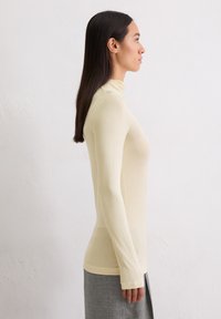 Vrouw met lang zwart haar, gekleed in een aansluitende crèmekleurige coltrui en een grijze rok, staande in profiel tegen een eenvoudige witte muur.