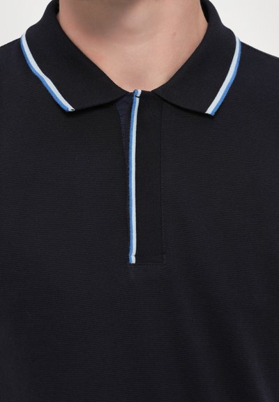 Pólo azul-marinho com um tecido texturizado, apresentando um colarinho e uma aba com riscas azul-claro nas bordas. Design minimalista.
