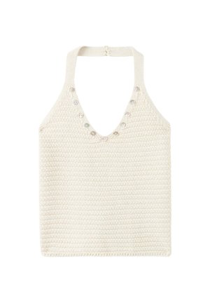 Jeune femme debout contre un mur beige, portant un débardeur en maille blanc, un short gris clair et des sandales marron cloutées.