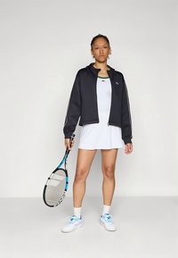 Μαύρο μπουφάν με φερμουάρ και λευκές ρίγες, συνδυασμένο με λευκό φόρεμα. Κρατά ρακέτα τένις και φοράει λευκά αθλητικά παπούτσια.