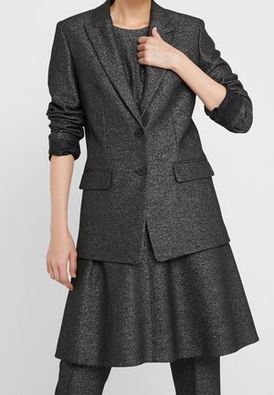 Kvinde iført en mørkegrå metallisk blazer med opkørte ærmer, matchende udvidet nederdel og bukser, hånd der justerer blazers revers.