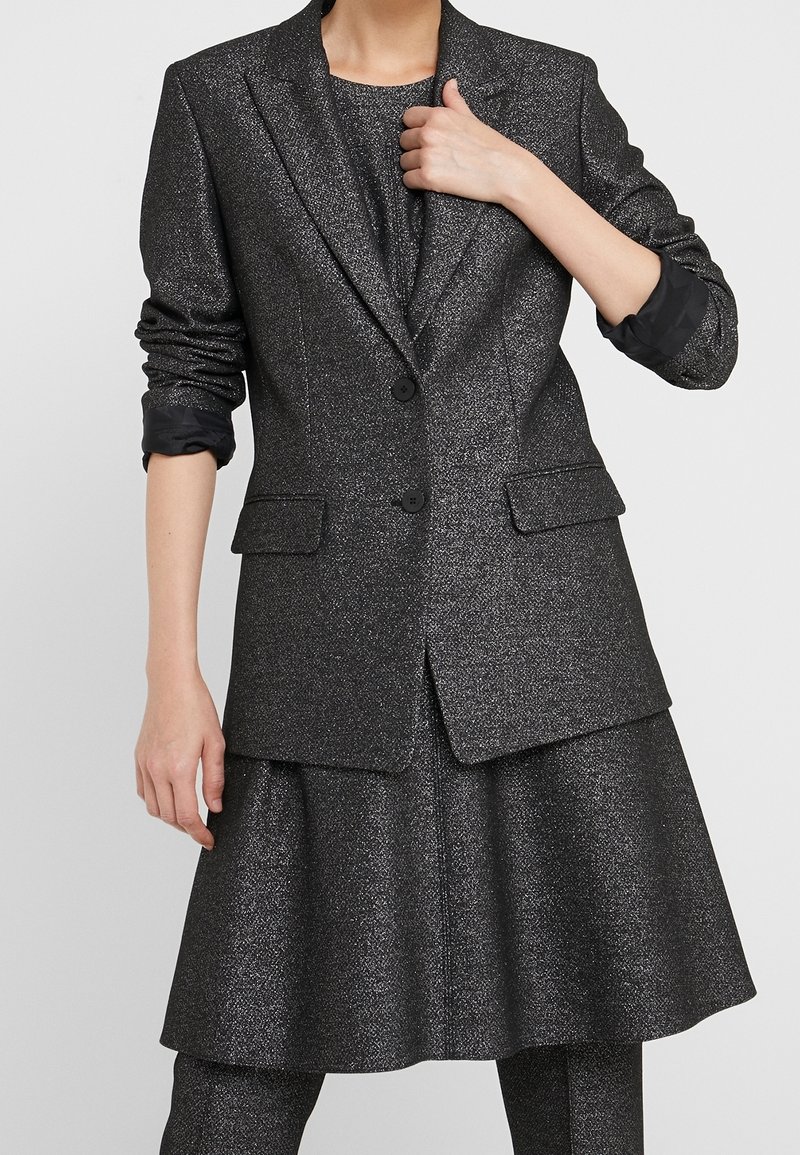 Femme portant un blazer métallique gris foncé avec manches retroussées, jupe évasée assortie et pantalon, main ajustant le revers du blazer.