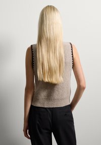 Beige ärmelloser Pullover mit gerippter Textur und dunklem Saum, kombiniert mit schwarzen Hosen; lange blonde Haare sind im Hintergrund sichtbar.