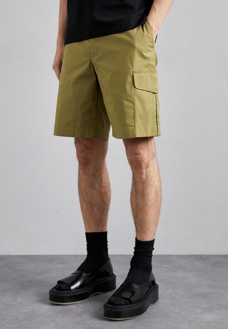 Personne portant un short cargo vert olive avec poche à rabat, des chaussettes noires et des sandales à plateforme noires à bout ouvert, debout sur un sol gris.