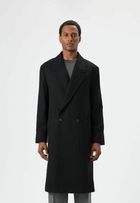 Zwarte wollen mantel met dubbele knopenrij en puntige revers, voorzien van twee knopen, lange mouwen en een losse pasvorm, gedragen over een grijze trui.