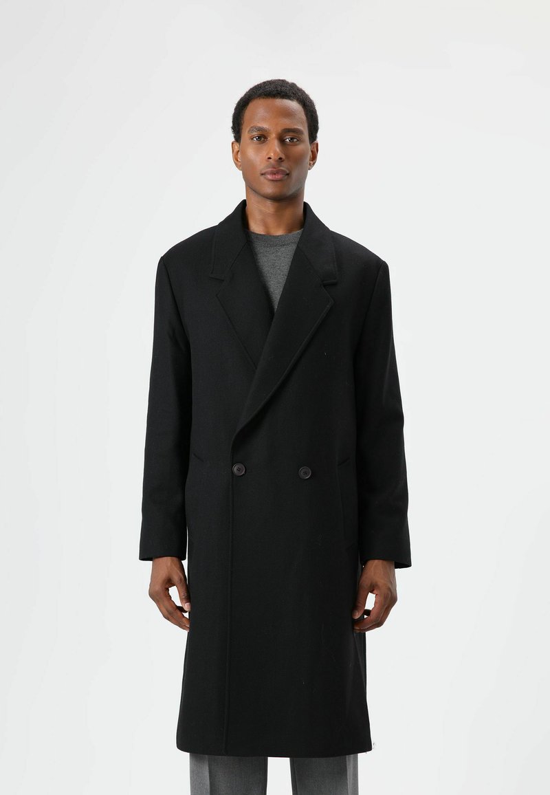 Zwarte wollen mantel met dubbele knopenrij en puntige revers, voorzien van twee knopen, lange mouwen en een losse pasvorm, gedragen over een grijze trui.