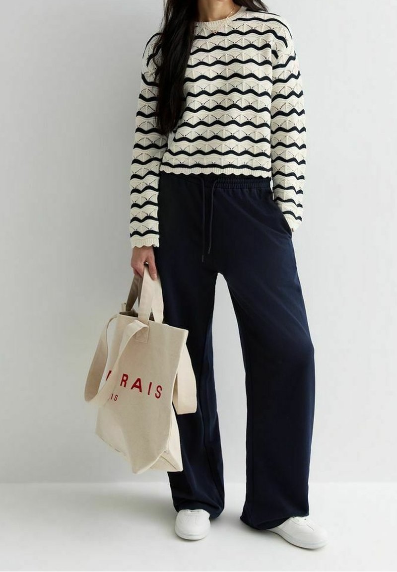 Personne portant un pull crème et noir à motif zigzag, pantalon ample bleu marine, baskets blanches, tenant un sac fourre-tout beige avec un texte rouge.
