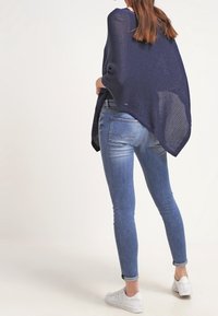 En marinblå stickad poncho hänger över en marinblå topp, kombinerad med ljusblå skinny jeans och vita sneakers, visad från baksidan.