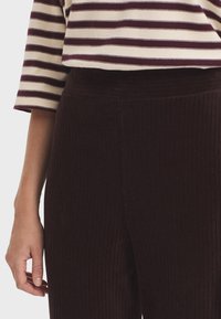 Person wearing dark brown ribbed pants and a beige shirt with maroon horizontal stripes, arm relaxed by their side.