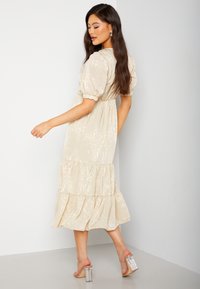 Beige gelaagde midi-jurk met puffmouwen, gemaakt van een textiel met structuur en een getailleerde taille. Gepaarde met doorzichtige haksandalen.