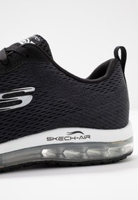 Svart idrottssko med meshuppbyggnad, vadderad krage och transparent luftkuddesula. Har en vit mellansula och "SKECH-AIR"-logotyp.