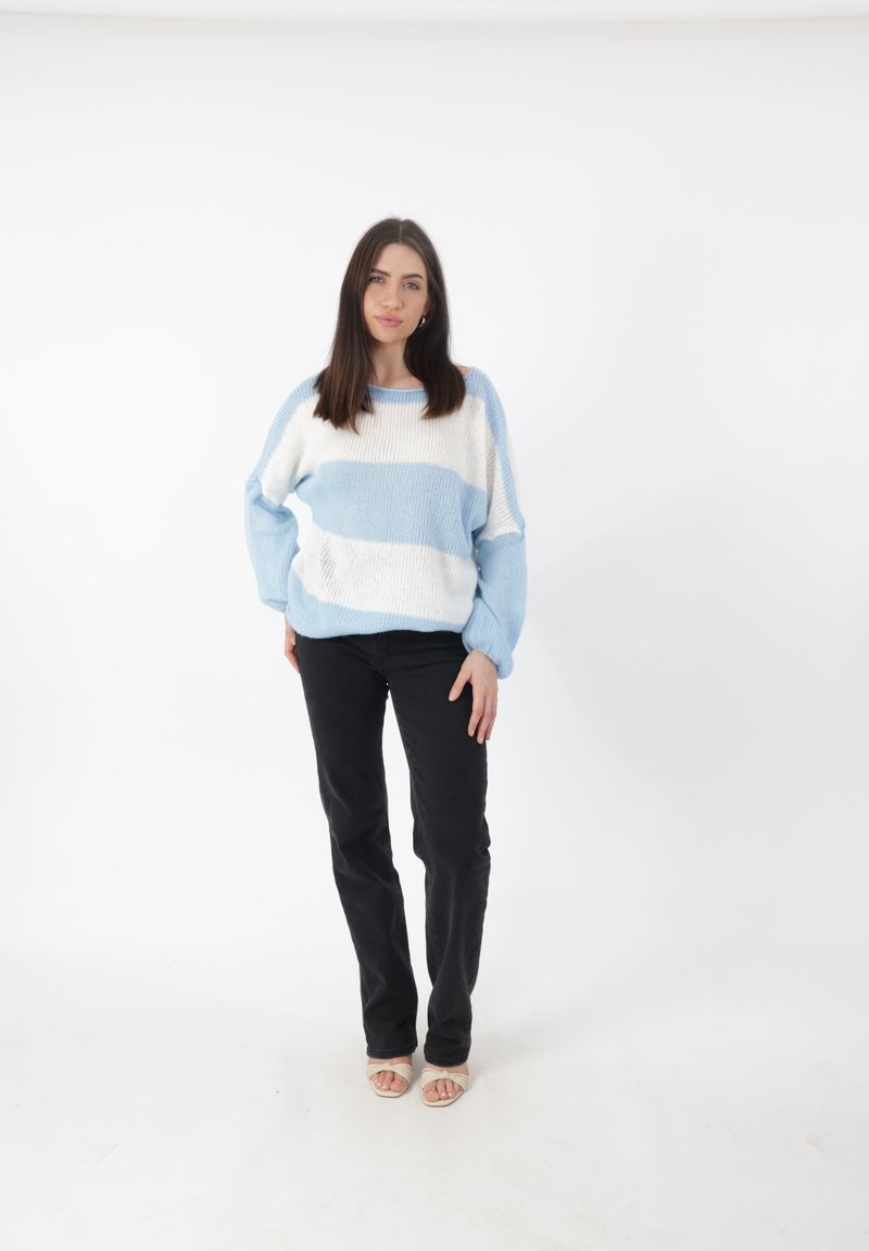 Hellblau und weiß gestreifter Strickpullover, übergroße Passform, kombiniert mit schwarzen, eng anliegenden Hosen und beigen Sandalen mit offenen Zehen. Einfarbiger weißer Hintergrund.