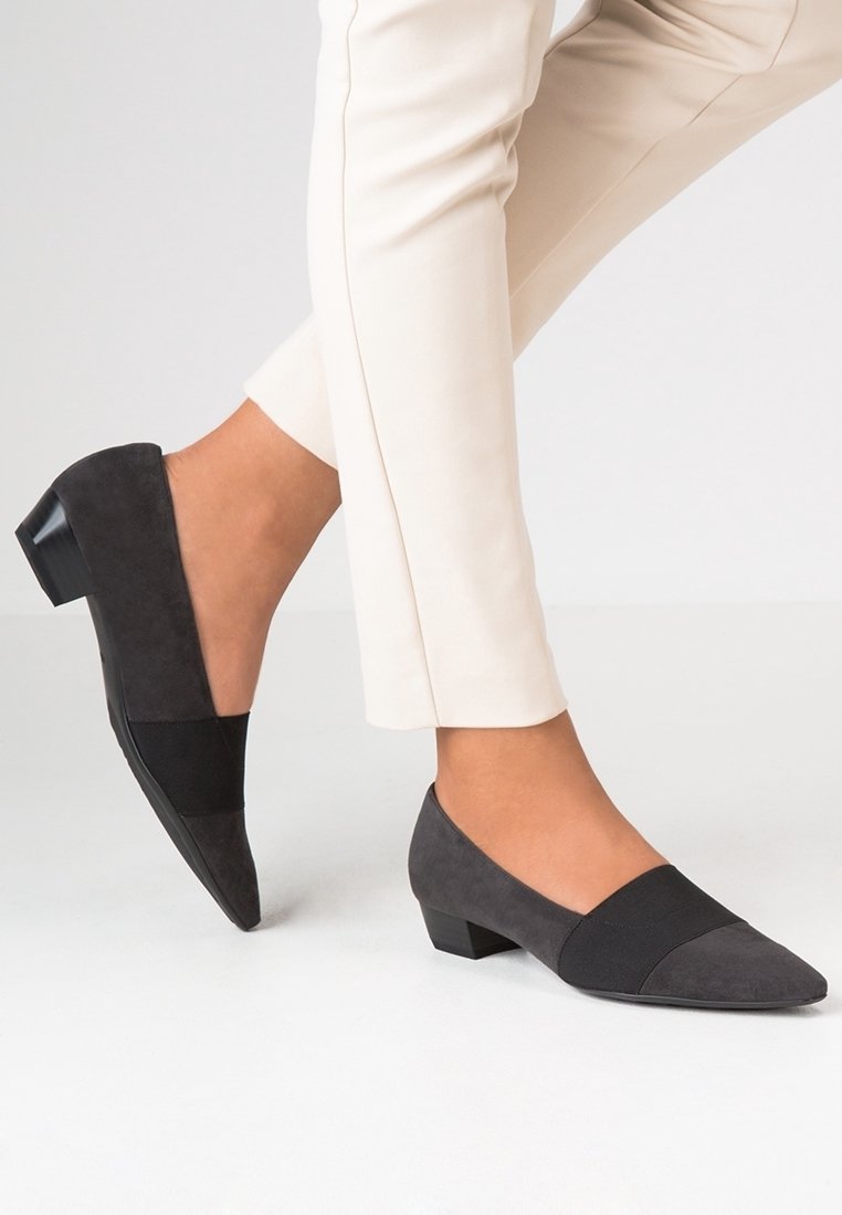 Femme portant des chaussures à talons bas pointues gris foncé et un pantalon slim beige clair, un pied légèrement levé du sol.