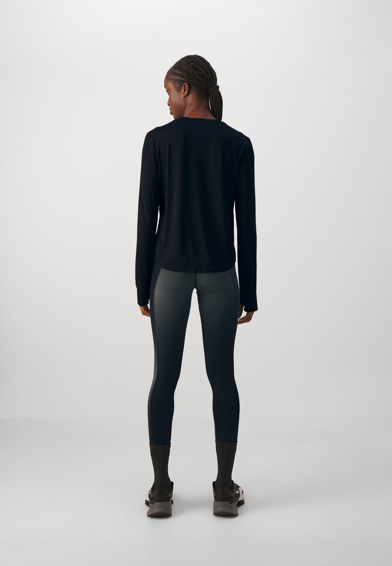 Top negro de manga larga con un corte relajado, combinado con leggings gris oscuro. El conjunto se complementa con calzado marrón. Fondo plano.