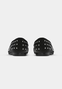 Sapatos pretos deslizantes com um upper suave, com pilares metálicos ao longo dos lados, uma sola plana e um design de bico arredondado.