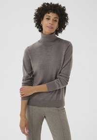 Brauner Rollkragenpullover aus weichem Strickstoff mit langen Ärmeln und geripptem Saum. Das Modell trägt ihn mit gemusterten Hosen.