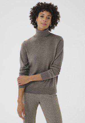 Pull à col roulé marron en maille douce, avec des manches longues et un ourlet côtelé. Le modèle le porte avec un pantalon à motifs.