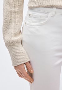 Άσπρο τζιν παντελόνι με λεπτή εφαρμογή, με μπροστινή τσέπη και λεπτομέρεια μεταλλικού κουμπιού, σε συνδυασμό με ανοιχτό μπεζ πλεκτό πουλόβερ.