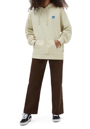 Personne portant un sweat à capuche beige avec une poche avant, un pantalon large marron et des baskets noir et blanc, debout les mains dans les poches.