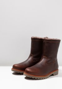 Bottes chevilles en cuir marron avec un col en fourrure douce, des coutures contrastées et une semelle robuste avec des motifs de grip. Fermeture éclair sur le côté.