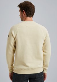 Sweatshirt crème avec un col rond côtelé et des poignets côtelés, présentant un patch logo discret sur la manche gauche, texture lisse et design décontracté.