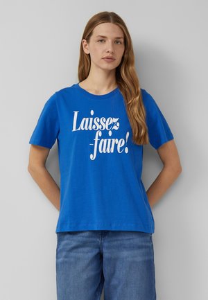 Jonge vrouw met lang haar, die een blauw T-shirt met de tekst "Laissez-faire!" draagt, staande tegen een effen lichtgrijze achtergrond.
