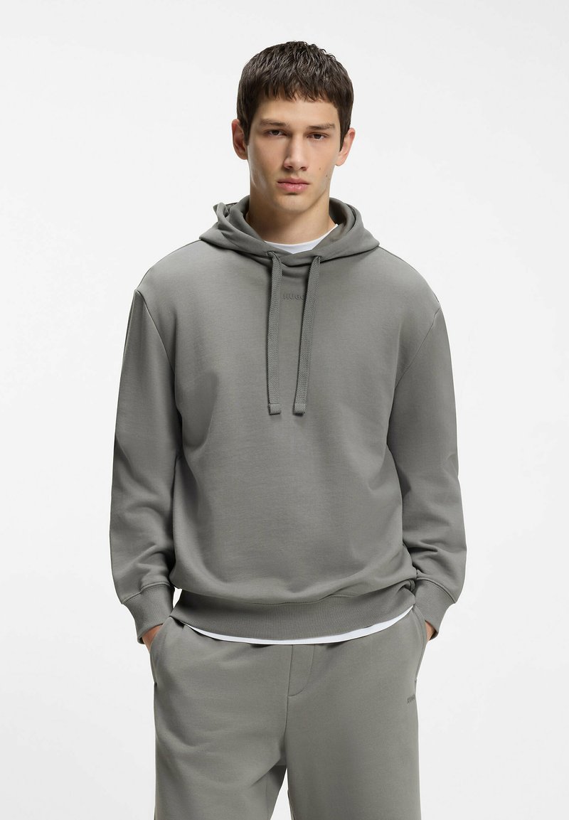 Jeune homme portant un sweat à capuche gris et un pantalon de survêtement assorti, les mains dans les poches, debout devant un fond blanc uni.