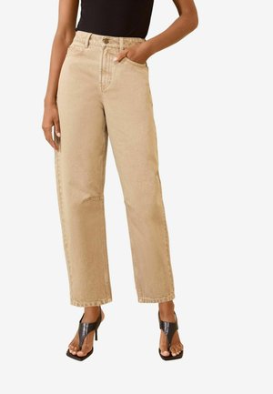 Vrouw draagt beige high-waisted wijde jeans en zwarte hoge hak thong sandalen, staat met één hand in de zak.