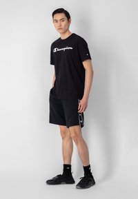 Camiseta de algodón negra con un logo blanco de "Champion", combinada con pantalones cortos negros que tienen acentos de logo en los lados. Los zapatos son negros con detalles texturizados.