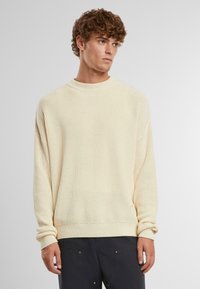 Pull en crème tricoté avec une texture côtelée, encolure ras du cou et coupe ample. Présente des manches longues avec des poignets repliés et un détail discret à l'ourlet.