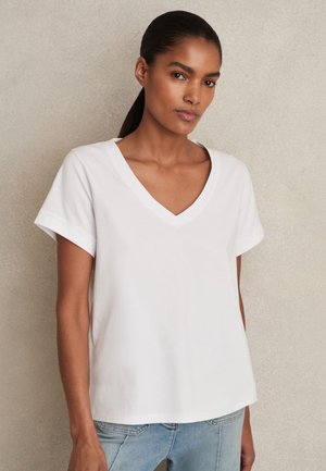 Sötét hajú nő lófarokkal, egyszerű fehér V-nyakú rövid ujjú pólót és világoskék farmert visel, bézs háttér előtt áll.