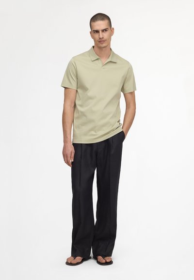 Man draagt een lichtgroen poloshirt met korte mouwen, losse zwarte broek en zwarte sandalen, staand met één hand in de zak tegen een witte achtergrond.